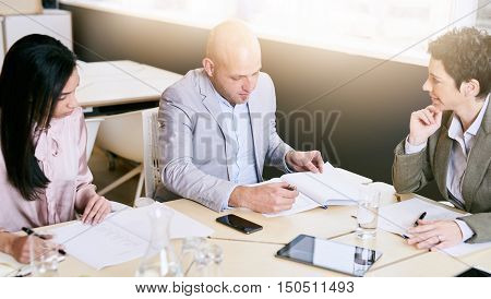 Business meeting between three executive employees, two female and one male, taking place in a birght conference table early in the morning to prepare for the week ahead.
