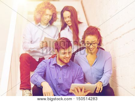 business, technology and startup concept - smiling creative team with tablet pc computer sitting on staircase