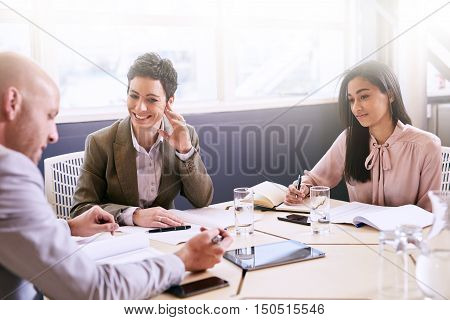 Business meeting between three executive employees, two female and one male, taking place in a birght conference table early in the morning to prepare for the week ahead.