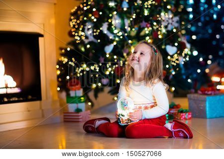 Family on Christmas eve at fireplace. Little girl opening Xmas presents holding snow globe. Child under Christmas tree with gift boxes. Decorated living room. Cozy winter evening at home.
