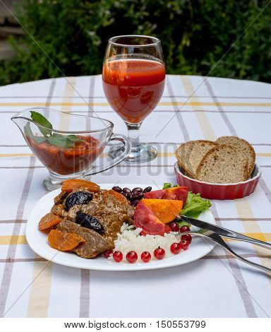 Beef stew with carrot and dried plum topping with rice beans and tomatoes. Served on a white plate with crusty bread home made sauce and glass of tomato juice.
