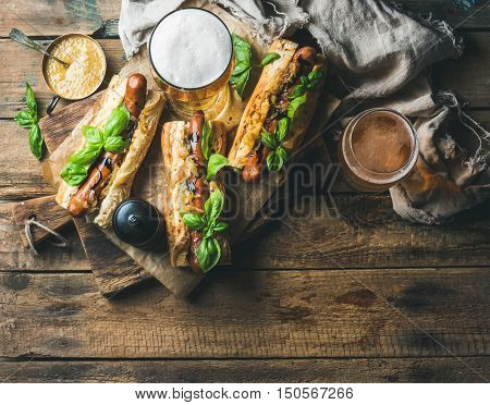 Glasses of wheat unfiltered beer and homemade grilled sausage dogs in baguette with mustard, caramelised onion and herbs on serving board over rustic wooden background, top view, copy space