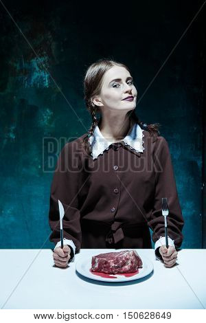 Portrait of a young girl with knife, fork and and a piece of raw meat on a plate. Girl in school uniform as killer. The image in the style of Halloween and Addams family