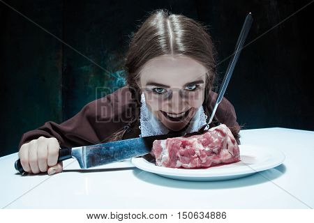 Portrait of a young girl with knife, fork and and a piece of raw meat on a plate. Girl in school uniform as killer. The image in the style of Halloween and Addams family