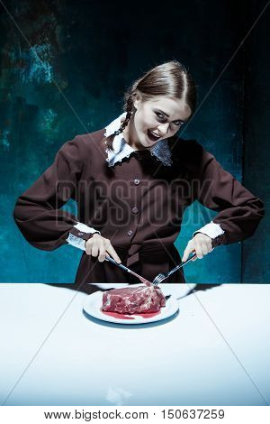 Portrait of a young girl with knife fork and and a piece of raw meat on a plate. Girl in school uniform as killer. The image in the style of Halloween and Addams family