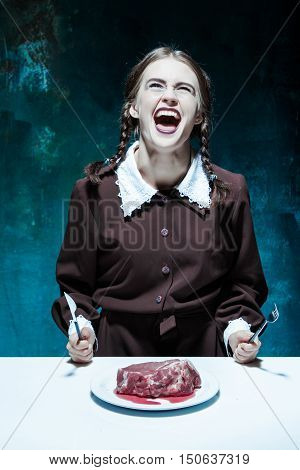 Portrait of a young girl with knife fork and and a piece of raw meat on a plate. Girl in school uniform as killer. The image in the style of Halloween and Addams family