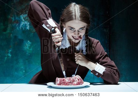 Portrait of a young girl with knife fork and and a piece of raw meat on a plate. Girl in school uniform as killer. The image in the style of Halloween and Addams family