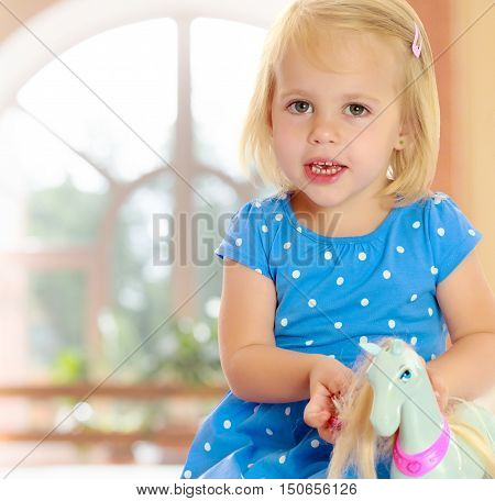 On the background of the hall with large semi-circular window.Cute little blonde girl playing with a toy horse. Girl wearing a blue dress with polka dots.