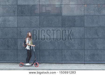 Joyful woman riding a kick scooter on modern fasade wall in background