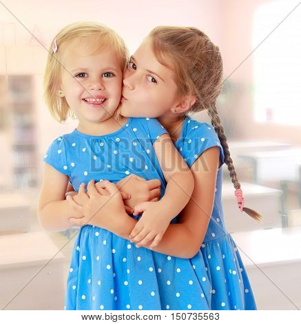 Two charming little girls, sisters , in identical blue dresses with polka dots. Older sister kissing the younger on the cheek.On the background of the school with desks