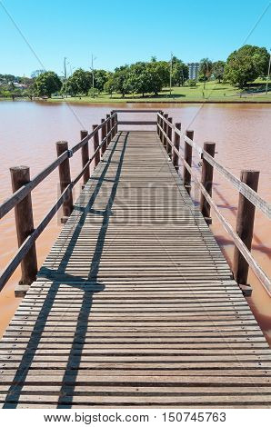 Deck made of wooden boards on lake water in a park with trees and green vegetation background on a sunny day. Rustic footbridge over water in perspective. Beautiful day in the park lake.