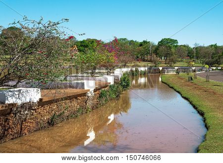 Stream of water between vegetation, some flowers and a blue sky in a park. Sunny day, reddish turbid water.