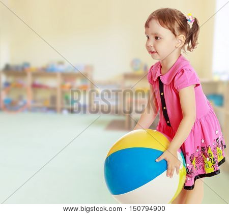 Beautiful little girl in a pink dress throwing a big striped ball. Turned sideways. Close-up.On blurred background the great hall of the kindergarten, with long racks where there are toys.