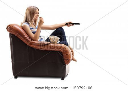 Young woman seated on an armchair changing channels on TV and eating popcorn isolated on white background