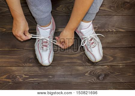 Tying Shoe laces girl sitting on the wooden floor. White sneakers on a dark wooden background.Side view.Fitness , sports.