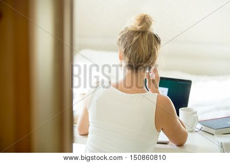 Woman sitting back talking on the phone at table with a cup and laptop on it. Rear view. Concept photo