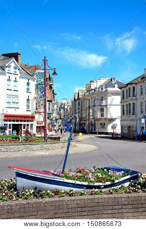 SEATON, UNITED KINGDOM - JULY 18, 2016 -Pretty flowerbeds on a traffic island with town centre buildings and shops to the rear and a fishing boat in the foreground Seaton Devon England UK Western Europe, July 18, 2016.