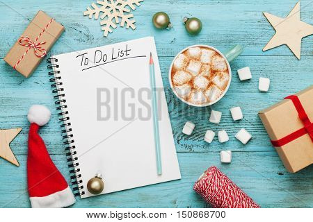 Cup of hot cocoa or chocolate with marshmallow holiday decorations and notebook with to do list on turquoise vintage table from above, christmas planning concept. Flat lay style.
