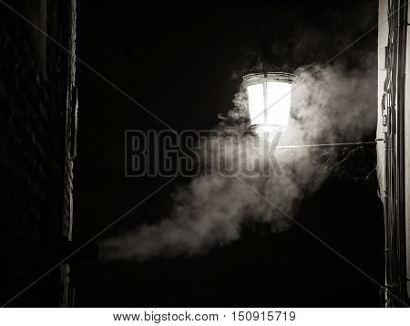 The smoke from a fireplace merges with the light of a street lamp during the night