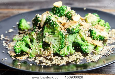 Close-up view of delicious steamed broccoli with garlic chips sprinkled with sesame seeds and dressed with soy sauce and ginger on the black plate. Tasty garnish dish of chinese cuisine. Green food