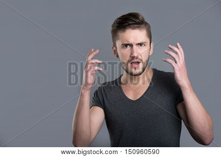 Shock. Puzzled bearded young man opening his mouth and raising his hands while standing isolated in grey background