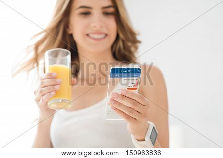 Technologies in mind. Joyful delighted smiling woman holding glass of juice and using smart phone while expressing positivity