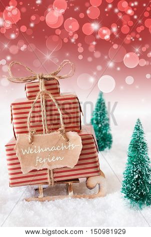 Vertical Image Of Sleigh Or Sled With Christmas Gifts Or Presents. Snowy Scenery With Snow And Trees. Red Sparkling Background With Bokeh. Label With German Text Weihnachtsferien Means Christmas Break