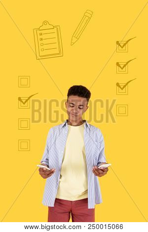Two Devices. Calm Attentive Modern Teenager Looking Attentively At Two Amazing Smartphones In His Ha