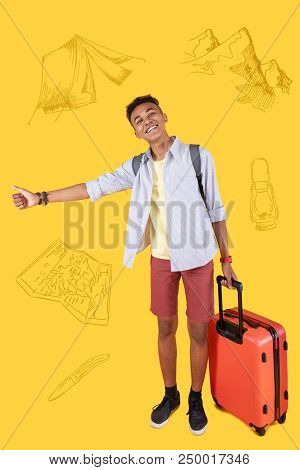 Hitchhiking Alone. Cheerful Experienced Hitchhiker Standing With His Thumb Up And Smiling While Tryi