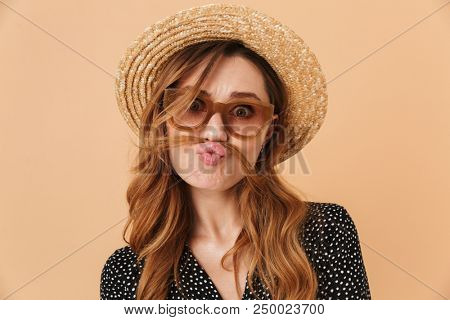 Portrait of amusing funny woman 20s wearing straw hat and sunglasses fooling around and putting hair like mustache isolated over beige background