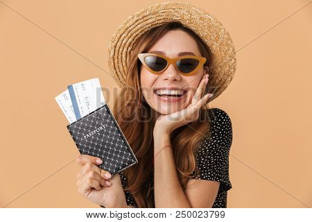 Charming brunette woman 20s wearing straw hat and sunglasses rejoicing while holding passport with travel tickets isolated over beige background