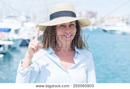 Beautiful middle age woman standing with the sea in the background surprised with an idea or question pointing finger with happy face, number one
