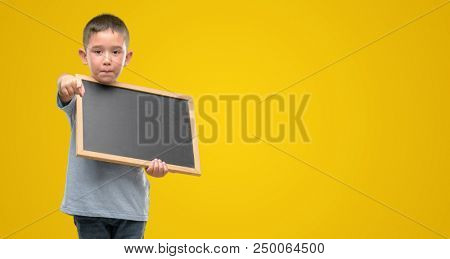 Dark haired little child holding a blackboard pointing with finger to the camera and to you, hand sign, positive and confident gesture from the front
