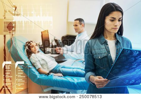Looking At Eeg Results. Calm Attentive Young Mother Looking At The Eeg Results Of Her Daughter While