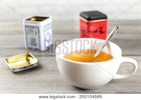 Cup Of Hot Black Tea, With Metal Loose Leaf Caddy In Back, On Gray Wood Desk.