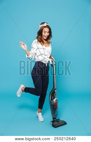 Full length portrait of european housewife 20s housekeeping and cleaning floor with vacuum cleaner isolated over blue background
