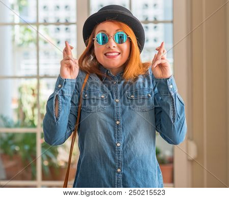 Stylish redhead woman wearing bowler hat and sunglasses smiling crossing fingers with hope and eyes closed. Luck and superstitious concept.