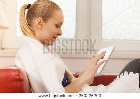 Young Woman Lying On Sofa Under Warm Blanket, Relaxing Using Tablet, Reading News And Checking Socia