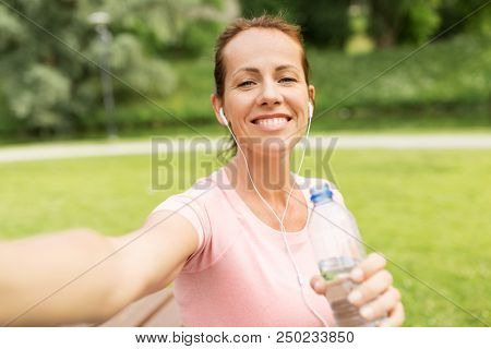 fitness, sport and healthy lifestyle concept - happy woman in earphones and water taking selfie in park