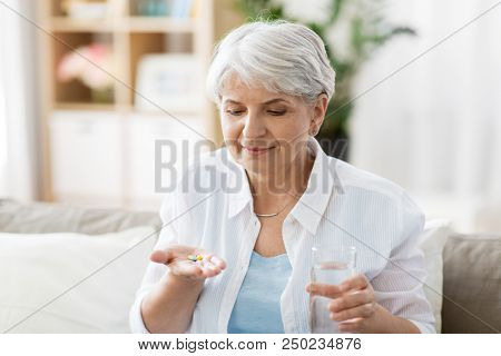 age, medicine, healthcare and people concept - senior woman with glass of water taking pills at home