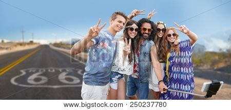 travel, technology and road trip concept - smiling young hippie friends taking picture by smartphone on selfie stick and showing peace hand sign over us route 66 background