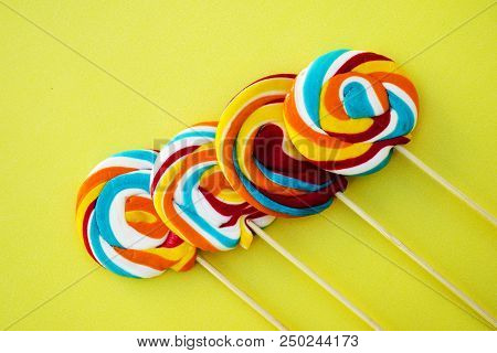 Colorful Hard Candy Lollipop On Yellow Pastel Background. Rainbow Sweet Pattern On Stick Flying Arou