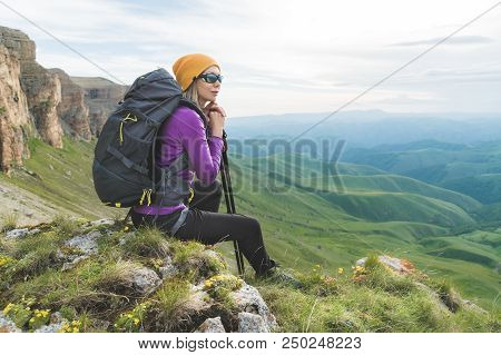 Smiling Girl Traveler In A Yellow Hat And A Pair Of Sunglasses Ssitting At The Foot Of Epic Rocks Wi