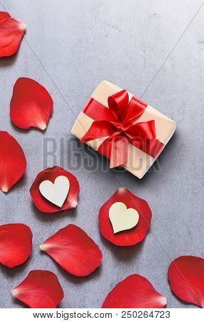 Gift In Craft Paper With Heart Decoration On Red Rose Petals Background On Grey Table. Romance Conce