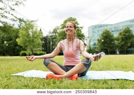 fitness, sport and technology concept - woman with smartphone and earphones meditating on yoga mat at park