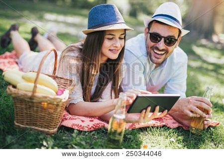 Couple Lying On A Picnic Blanket In A Park With A Picnic Basket Filled With Fruit, They Are Using Di