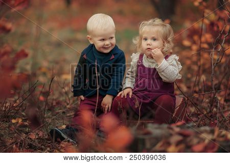 Little Cute Girl And Boy Blondes Are Sitting On Leaves In Autumn Forest. Romantic Couple