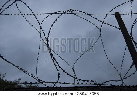 Barbed Wire At The Top Of The Fence Against The Background Of A Dramatic Sky. Without Color