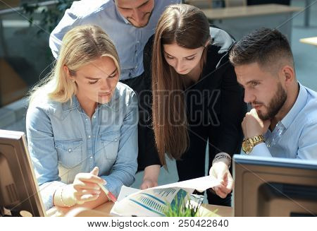 Group Of Young People In Casual Wear Sitting At The Office Desk And Discussing Something While Looki