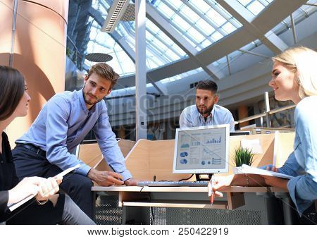 Group Of Young People In Casual Wear Sitting At The Office Desk And Discussing Something While Looki